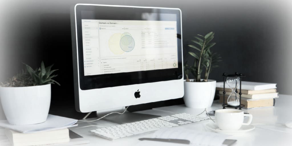 office desktop with computer screen displaying company financials, surrounded by plants, coffee cup, and stacks of books