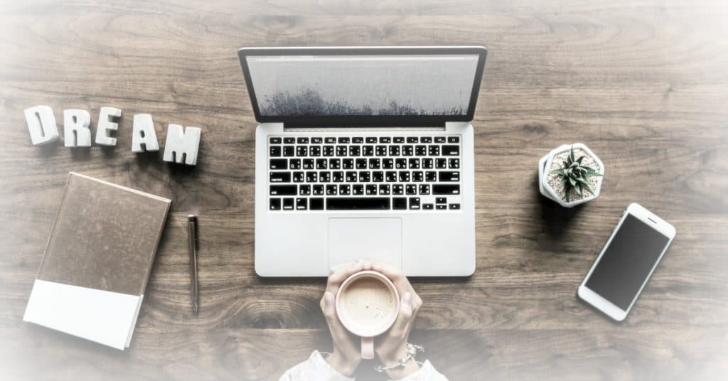 laptop, notepad and pen, mug of coffee, and cell phone on a desk