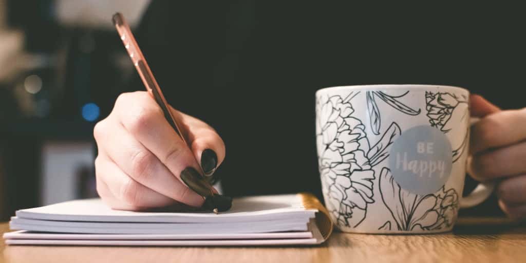 woman writing a financial mission statement while drinking from a mug that says be happy on it