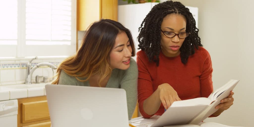 two women looking at a book with a laptop by their side