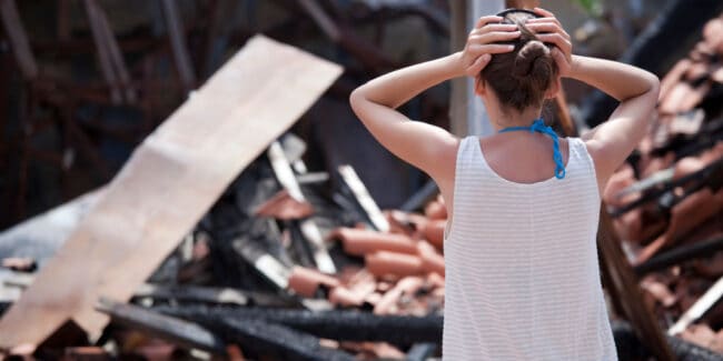 Woman standing in front of burned out house with both hands on her head