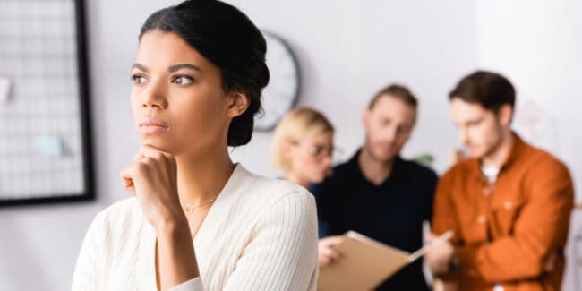 pensive african american in the workplace with group of other employees conversing behind her