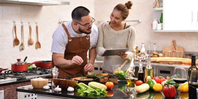 Loving young Asian couple using digital tablet and meal prepping in kitchen making healthy food together feeling fun