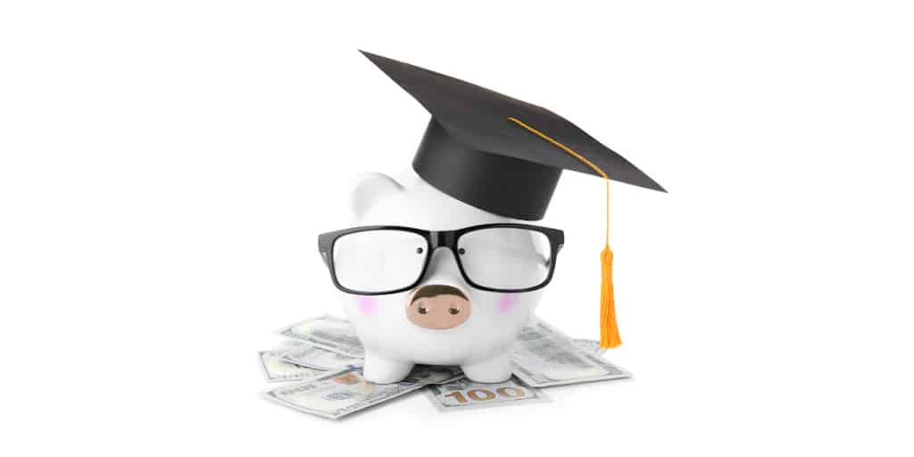 white piggy bank wearing black graduation cap and glasses sitting on stack of cash money