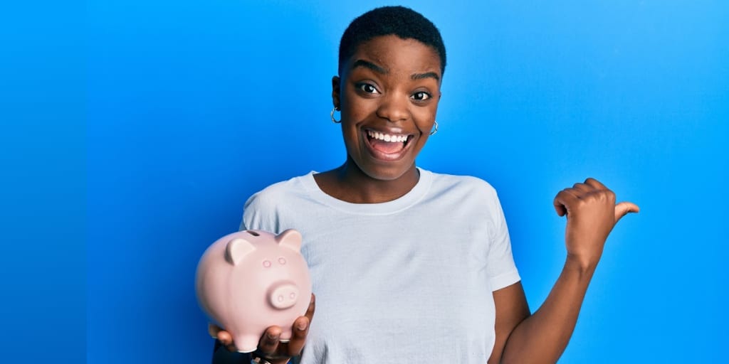 Young african american woman holding piggy bank pointing thumb up to the side while smiling wide