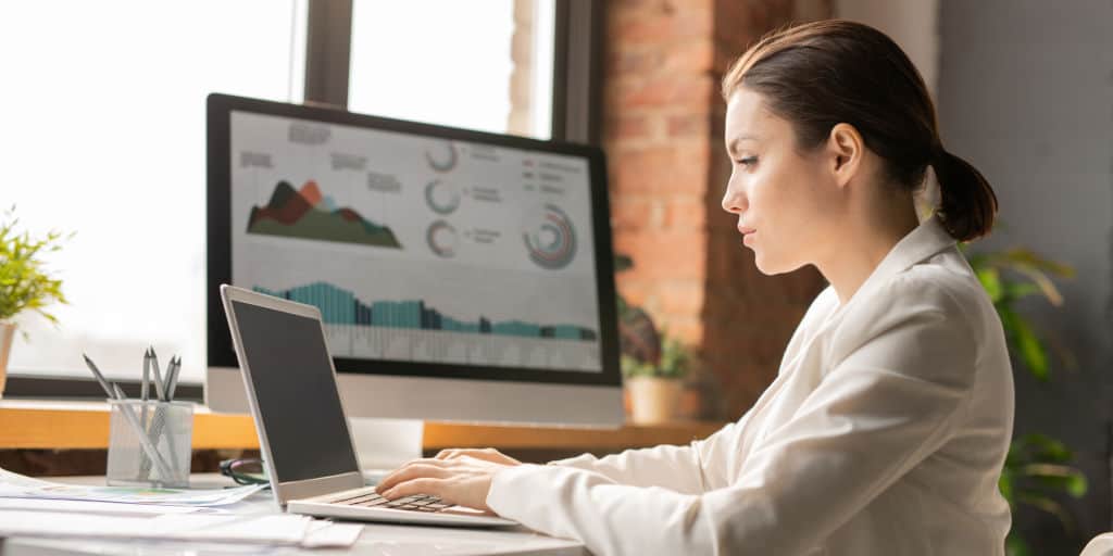 female investor at desk using laptop computer attached to large monitor