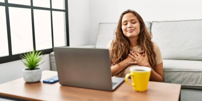 hispanic woman using computer laptop at home smiling with hands on chest with closed eyes and grateful gesture on face. financial health concept.