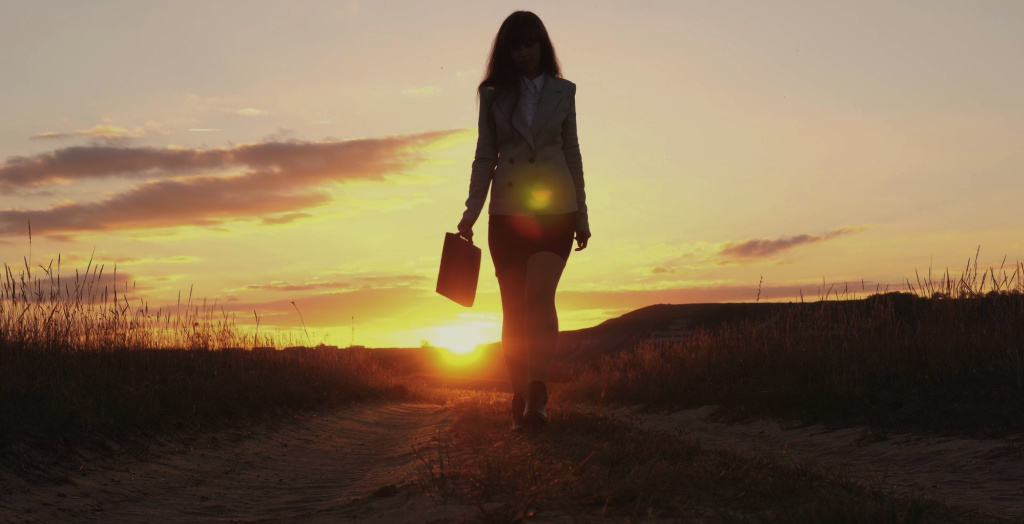 silhouette of business woman walking on country road carrying a briefcase with sunset glowing behind her