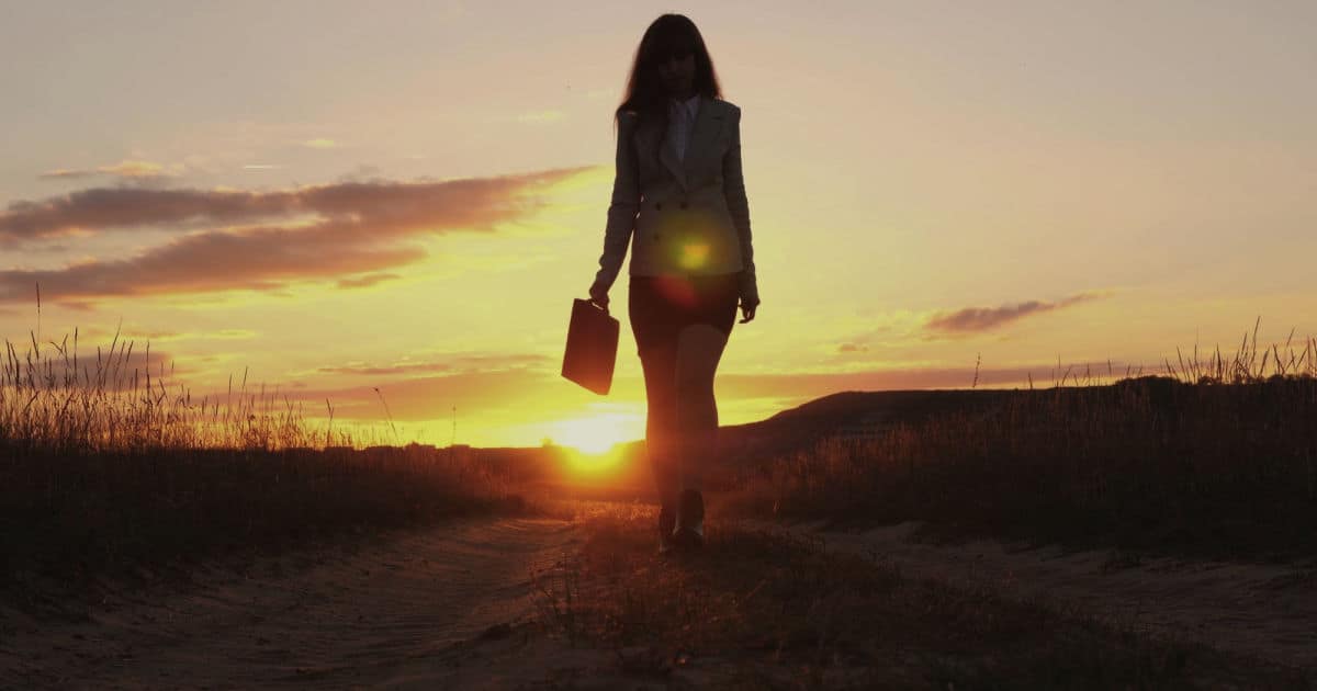silhouette of business woman walking on country road carrying a briefcase with sunset glowing behind her