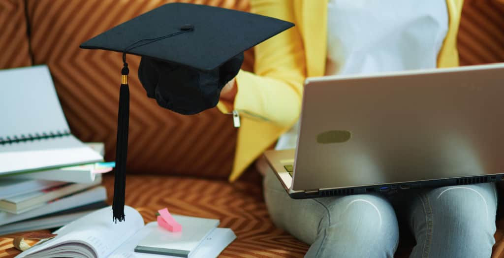 college student in jeans and yellow jacket sitting on couch with laptop and text books holding a graduation cap 
