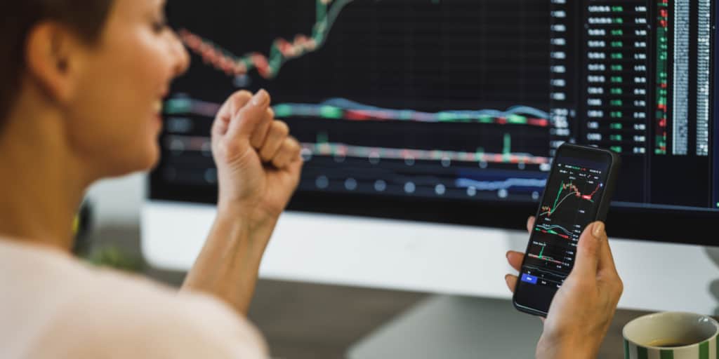 woman celebrating for success while day trading with her smartphone and computer at home