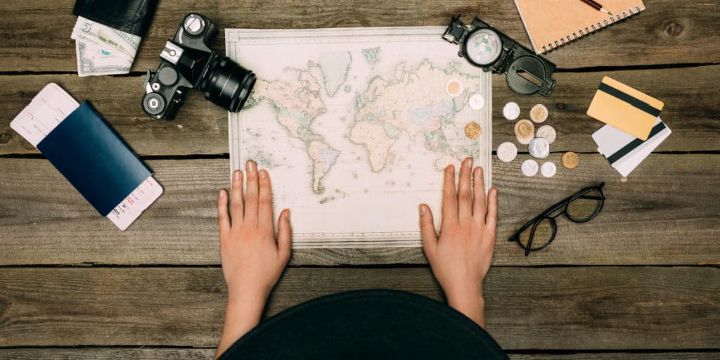 travel map on wood desk top with travel accessories including passport, camera, currency, a journal, and eyeglasses