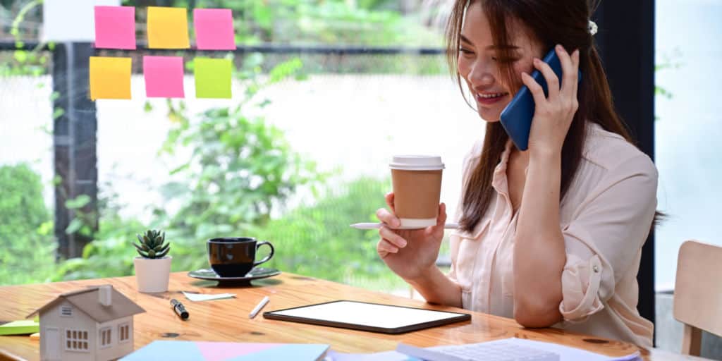 Smiling young female drinking coffee and talking on mobile phone at office doing due diligence on real estate property.