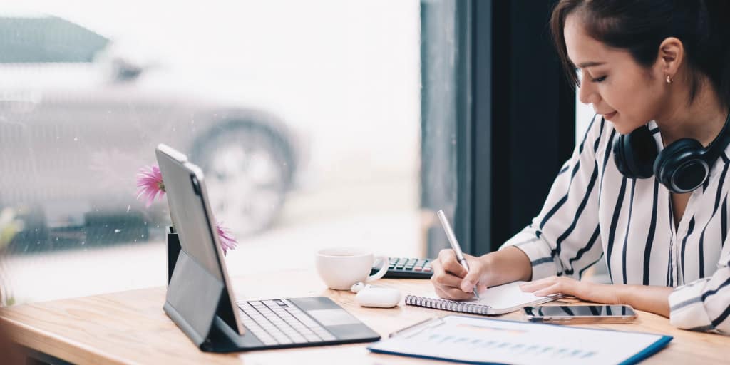 Asian woman wearing headphones listening to a personal finance podcast on laptop and taking notes