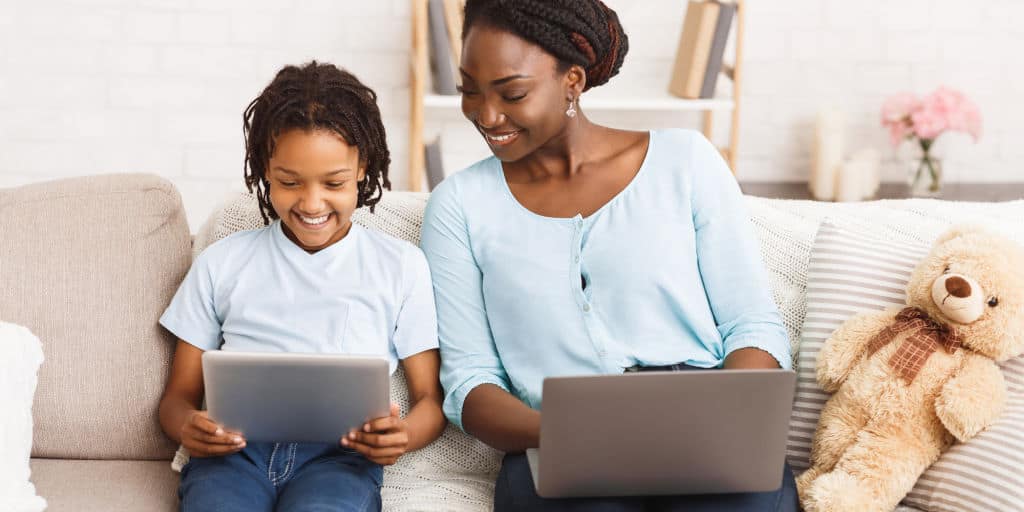 african american mom and daughter using laptop and tablet while sitting on couch