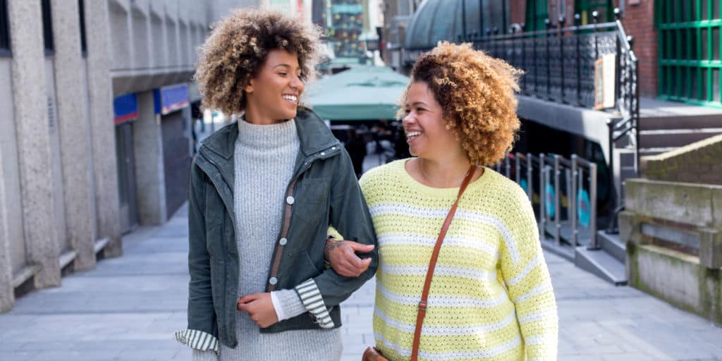 Sisters walking arm-in-arm through the city. They are looking at each other and laughing.