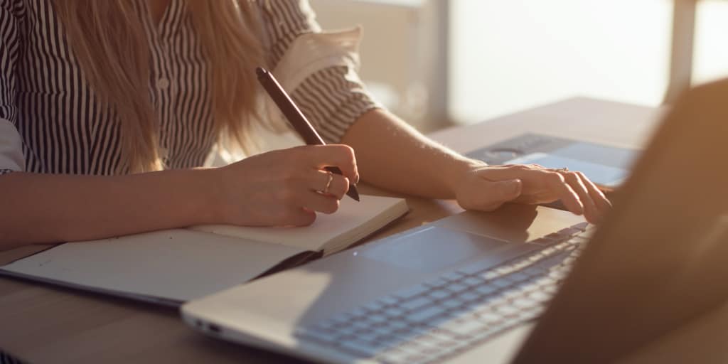 Manos femeninas con la escritura de bolígrafo en el cuaderno y el uso de la computadora portátil